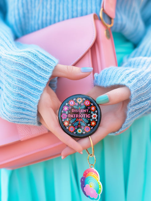 Round black pinback button with vibrant floral embroidery design surrounding bold pink and red text reading "Dissent is Patriotic."